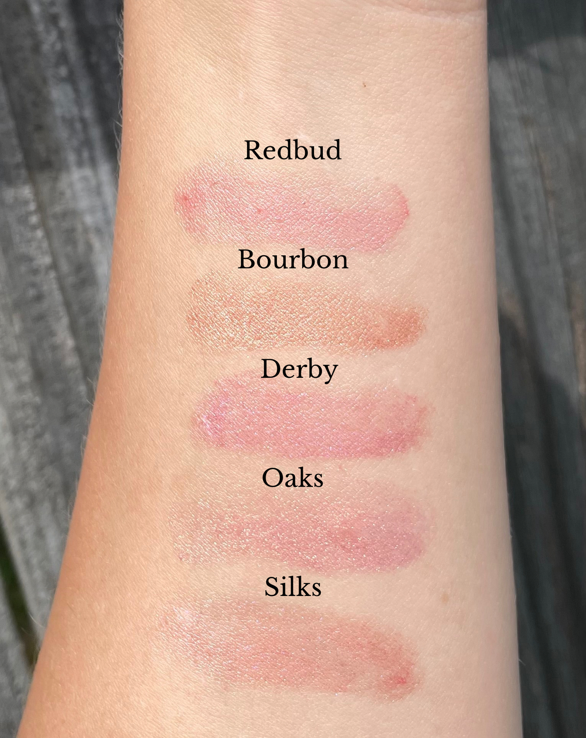 A close-up of an arm showing five makeup swatches labeled Redbud, Bourbon, Derby, Oaks, and Silks, with different pink and peachy-rose shades applied in horizontal strokes.
